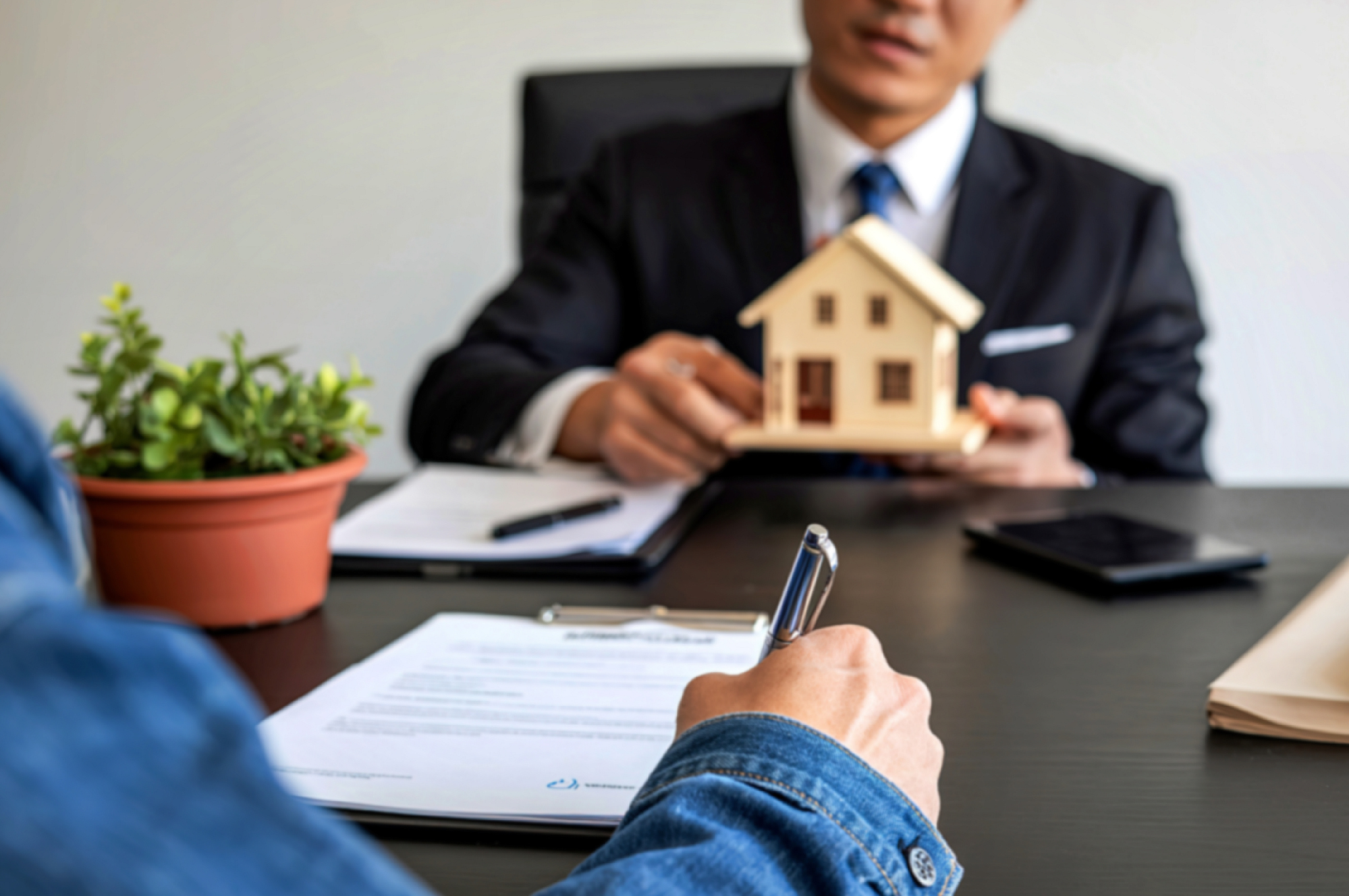 A client signing real estate documents across a desk from a realtor holding a miniature house model, representing a home sale or purchase transaction.